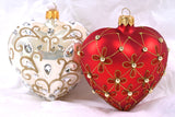 To hjerteformede julekuler i malt glass. Til venstre: hvitt hjerte dekorert me hullmønster og blanke glassteiner formet som dråper. Til høyre: rødt hjerte i glass med blomsterdekor og gullperler. Julepynt